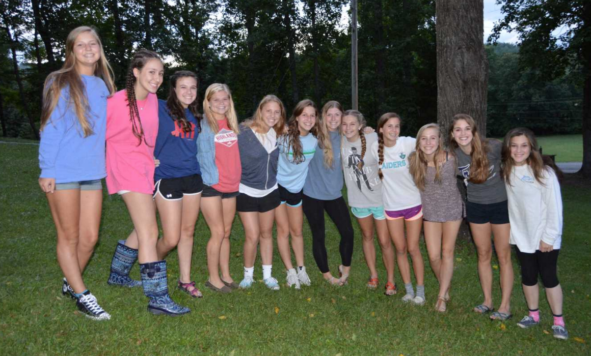 Group of twelve girls standing arm in arm on a grassy lawn during a senior trip, with trees in the background